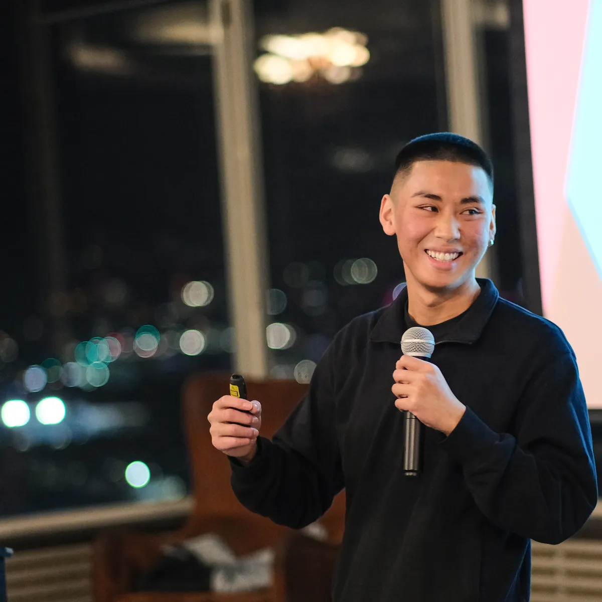 Jordan Pham smiling on stage with a microphone, Melbourne city lights through the windows behind him