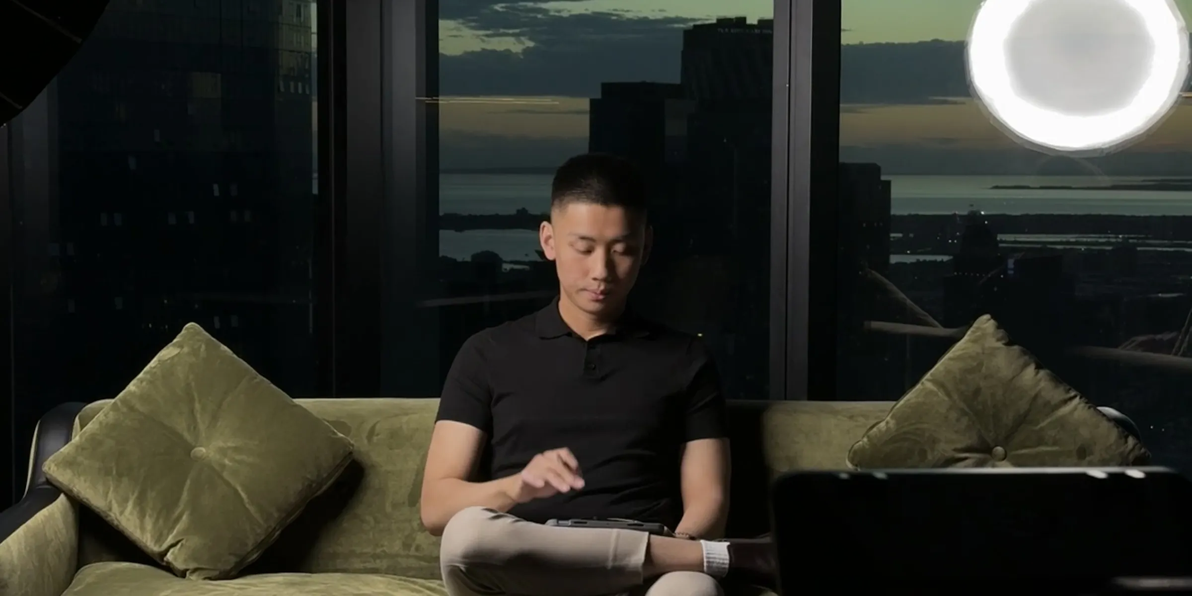 Jordan Pham on a green velvet sofa between takes, lights and camera set up, Melbourne skyline at dusk behind him